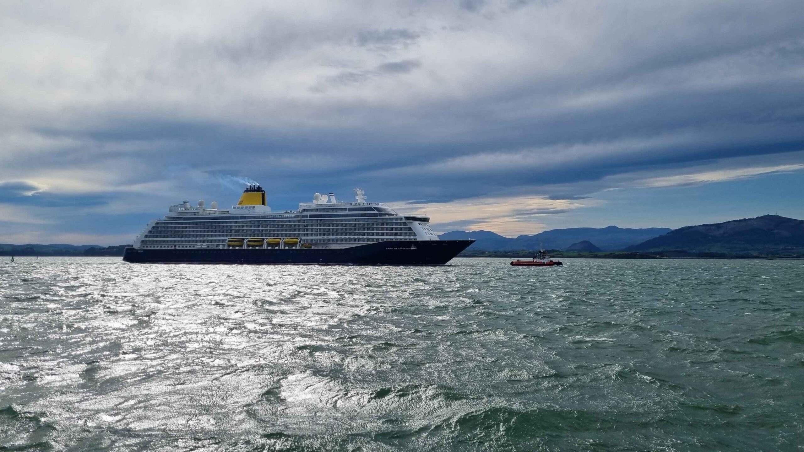 Spirit of Adventure Cruise Ship in the Bay of SantanderThe image shows the impressive cruise ship "Spirit of Adventure" sailing in the picturesque Bay of Santander. This majestic vessel, painted in elegant blue and white colors with yellow accents, stands out in the bay's silvery water, reflecting the sunlight on a partly cloudy day. In the scene, a small tugboat escorts the cruise ship, providing an interesting contrast with the calm mountains rising on the horizon. The Bay of Santander, known for its natural beauty and strategic location, offers a spectacular setting for cruise navigation. The light of dawn or dusk adds a special shine to the water, creating a visual effect that highlights the grandeur of the "Spirit of Adventure." This corner of Cantabria is not only a key point for maritime tourism but also an attractive destination for lovers of coastal landscapes and navigation.